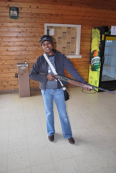 Person in a beanie and jeans holds a shotgun, standing indoors with wood-paneled walls