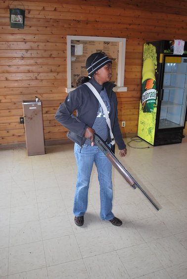 Woman in a beanie and jacket holding a shotgun, standing indoors near a Gatorade cooler