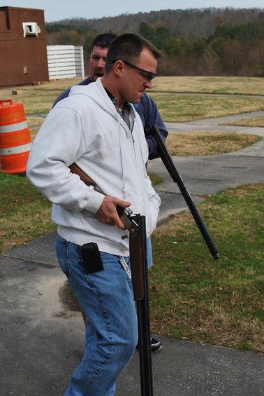 Two men stand at a shooting range, one holding a shotgun