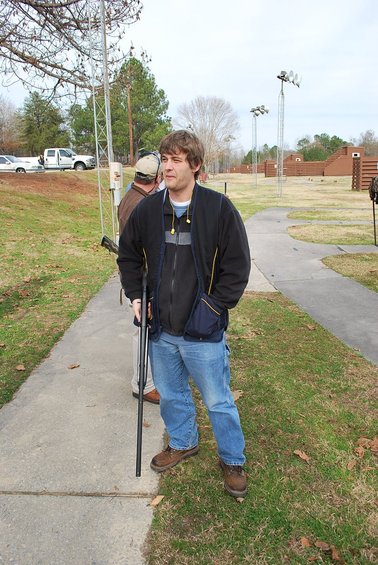 Man holding a shotgun at a shooting range, with other people and a white truck in the background