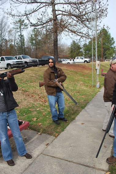 Several pickup trucks parked on a grassy area near a shooting range with people holding shotguns