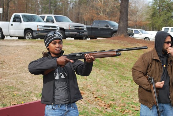 Woman in black jacket and beanie aims a shotgun; man in brown jacket stands nearby. Several pickup trucks are parked in the background