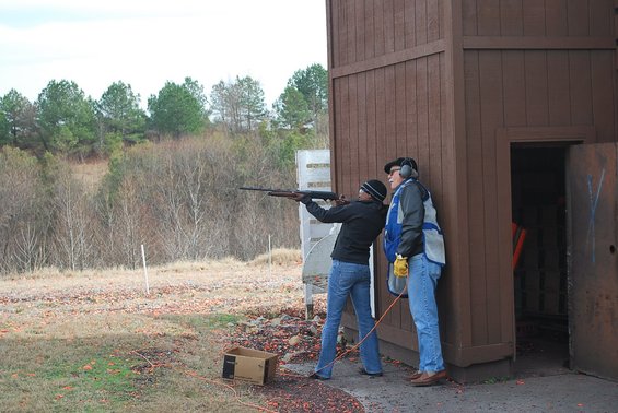 Two people at a shooting range, one aiming a shotgun with an instructor beside them