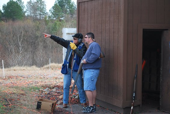 Man in blue vest points, man in blue sweatshirt holds shotgun at shooting range
