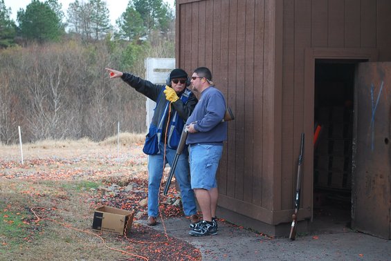 Two men stand near a wooden structure at a shooting range, one pointing into the distance