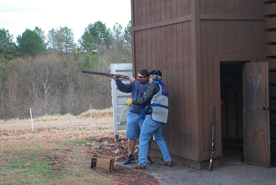 Two men at a shooting range, one aiming a shotgun, with a wooden structure and trees in the background