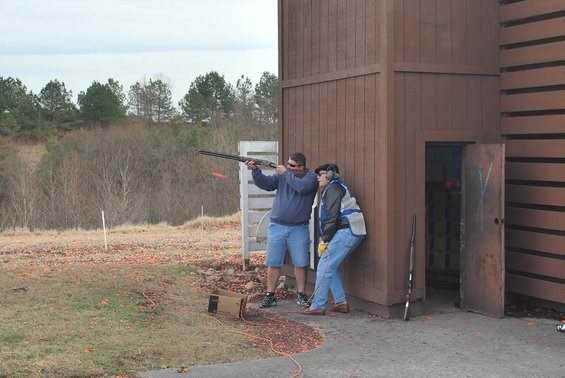 Two men at a clay pigeon shooting range, one firing a shotgun