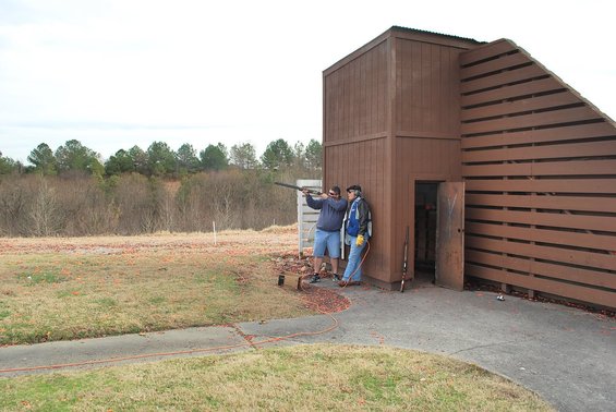 Two guys at a trap shooting range, one shooting, one watching. There's a wooden structure behind them