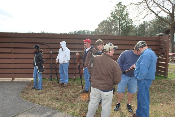Group of men and one woman standing near a wooden fence with shotguns