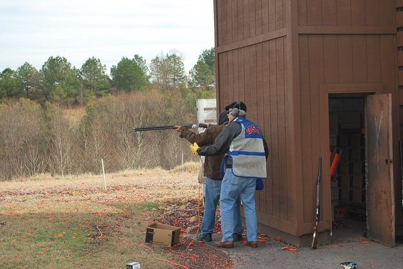 Two people at a shooting range, one aiming a shotgun. A wooden structure is to the right