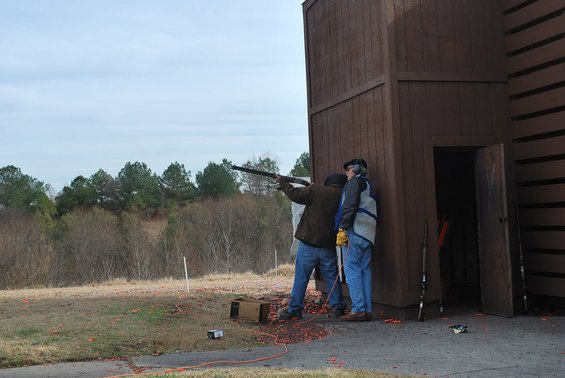 Two men at a shooting range, one aiming a shotgun, near a wooden structure