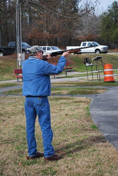 Man in blue jacket and jeans aims a shotgun at a shooting range; two trucks are parked in the background