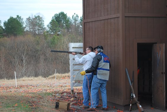 Two men at a clay shooting range, one aiming a shotgun