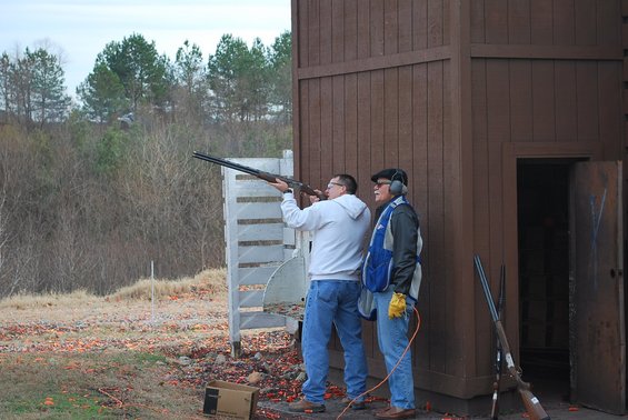 Two men stand at a clay shooting range, one aiming a shotgun