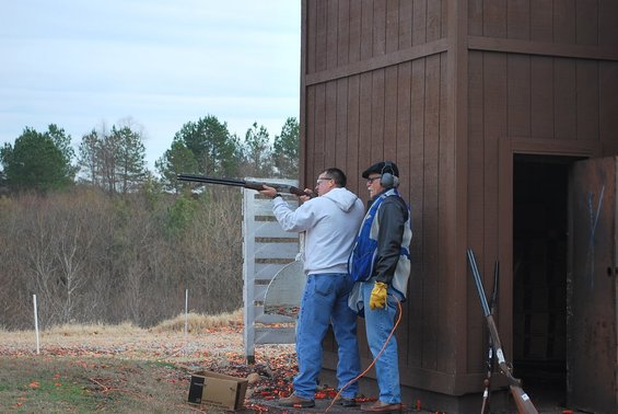 Two men at a shooting range, one aiming a shotgun