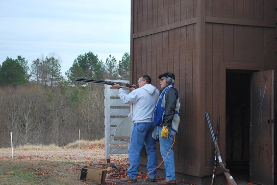Man in white hoodie aims shotgun while another man watches at outdoor shooting range