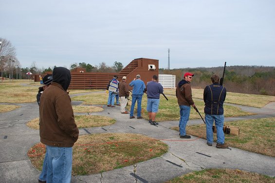 People stand in a line at a shooting range, holding shotguns. A wooden structure is in the background