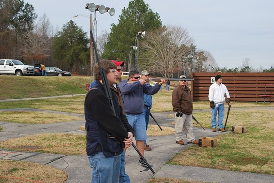 People with shotguns at a shooting range, with a white pickup truck and a car parked on a hill in the background
