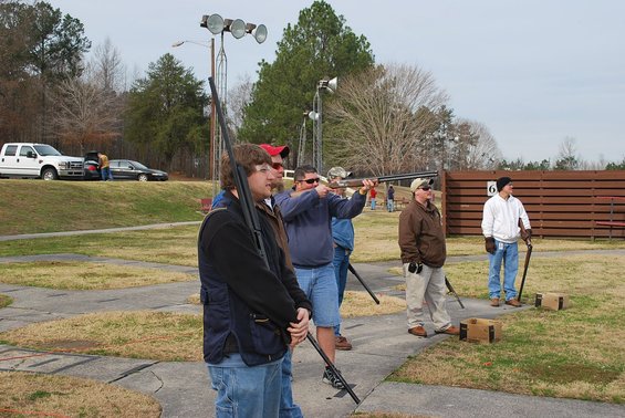 Guys with shotguns at a shooting range, one guy is shooting. White pickup truck and a car parked in the background