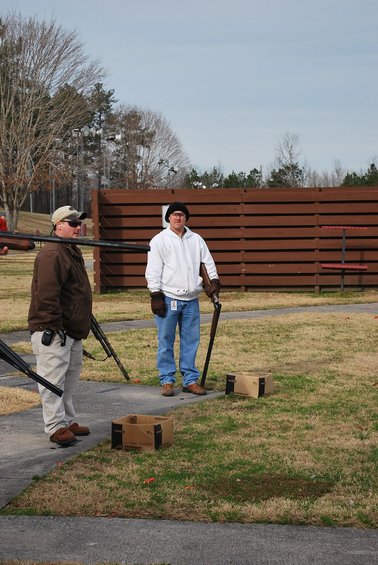 Two men stand at a shooting range with shotguns and ammunition boxes