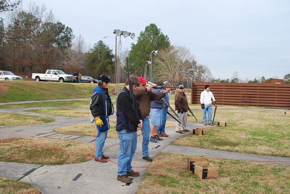 Skeet shooting range with several people taking aim and others observing. Trucks are parked in the background