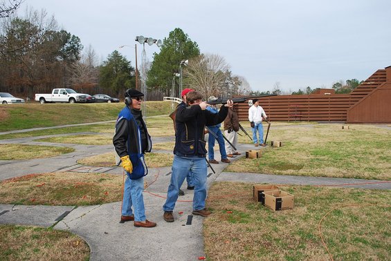 People at a shooting range with clay targets being launched; a white pickup truck is parked in the background