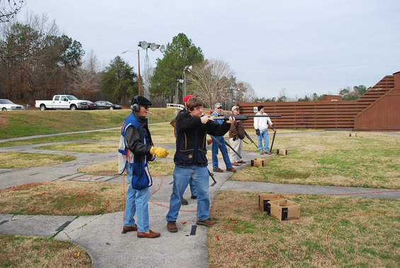 Guys at a shooting range, one guy is taking a shot with a shotgun. Another guy is watching him. There are cars parked in the background