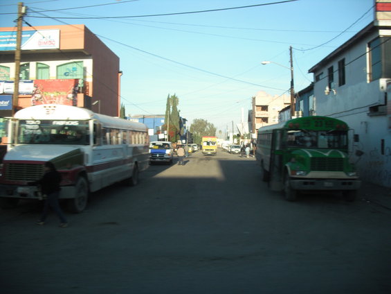 Street with two buses, one white and red, the other green. Other vehicles and buildings visible