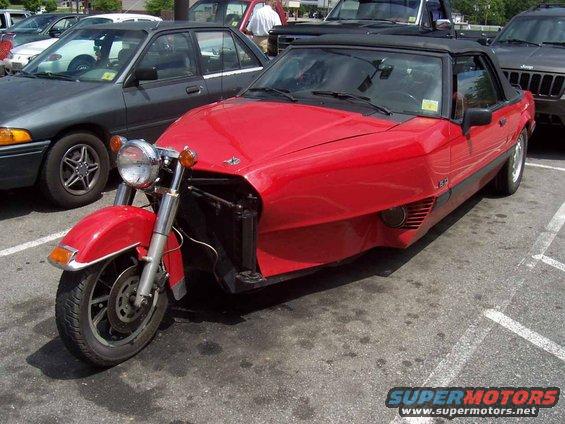 Red custom trike with motorcycle front, convertible top, and car body parked in a lot
