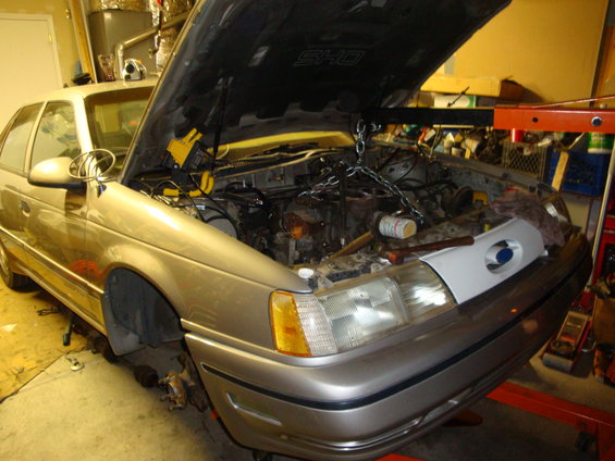 1990 Ford Taurus SHO engine bay with engine hoist attached, hood open, front wheels removed