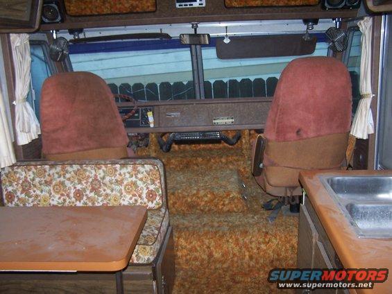 RV interior with brown seats, floral upholstery, and orange shag carpet. A table is visible on the left