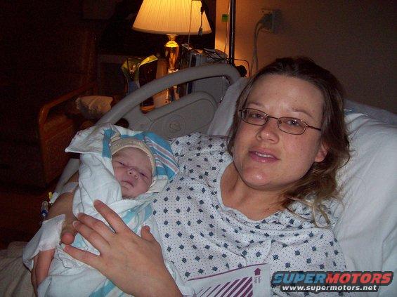 Woman with glasses holding a swaddled newborn baby in a hospital bed, with a lamp and medical equipment visible
