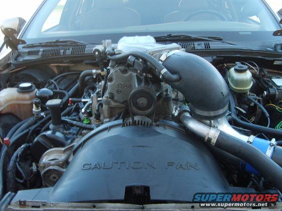 Engine bay with a supercharger and large fan shroud. Text on shroud says 'CAUTION FAN'