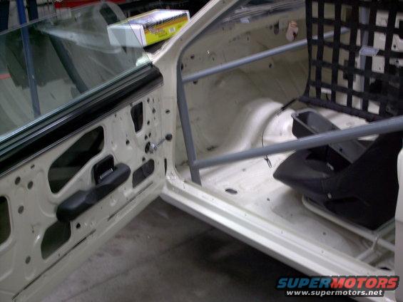 Car interior with roll cage, racing seat, and stripped door panel. White exterior
