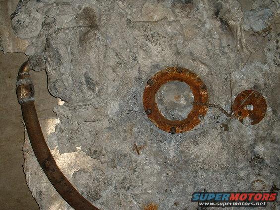 Rusted pipe fitting with a chain attached to a small circular plate, mounted on a rough concrete surface