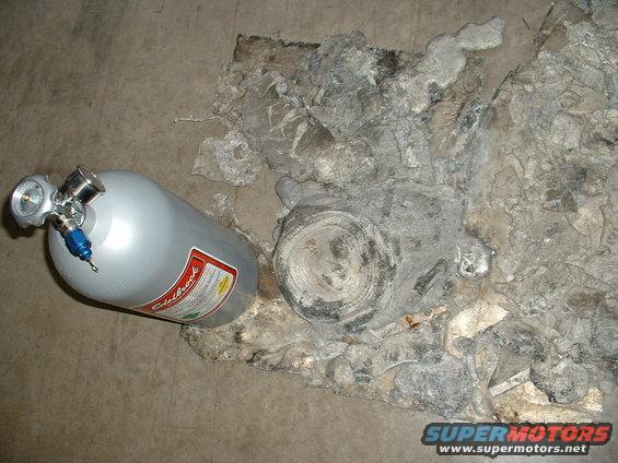 Silver Edelbrock nitrous oxide bottle next to a damaged engine block on a concrete floor