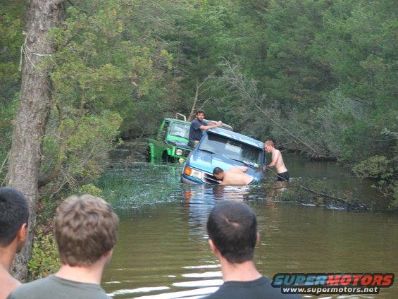 people trying to push a blue SUV and a green off-road vehicle out of the water