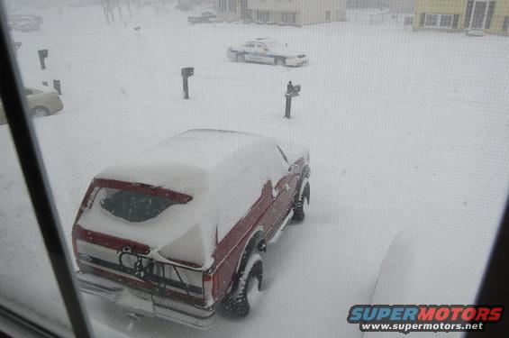 Red pickup truck covered in snow, parked in a driveway during a blizzard. Another car is visible in the background
