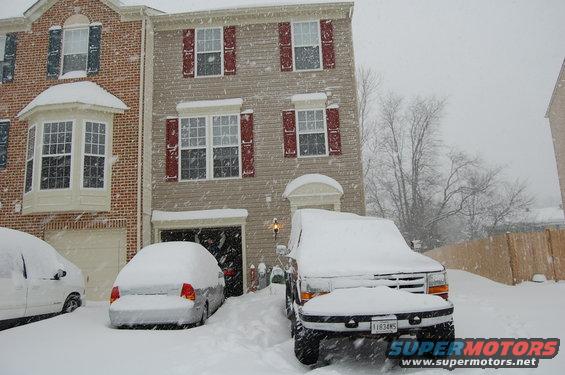 Two cars covered in snow in front of a house during a snowstorm