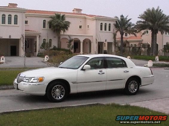 white 2002 Lincoln Town Car is parked on the street in front of a large house with palm trees