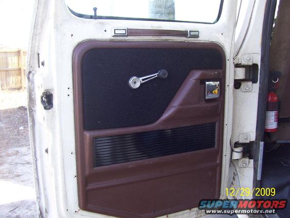 White vehicle interior door panel with brown trim, black fabric insert, and chrome window crank. Fire extinguisher visible
