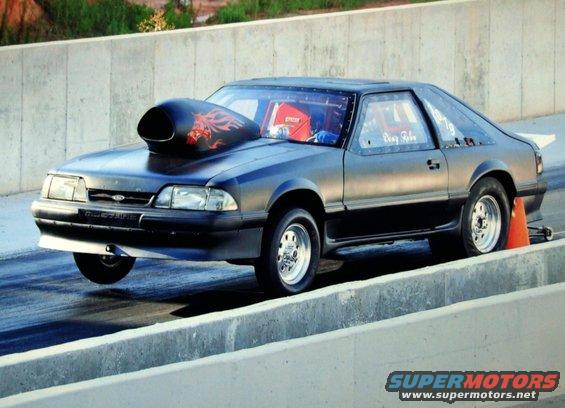 Black 1990 Ford Mustang drag car with a large hood scoop, racing down a track