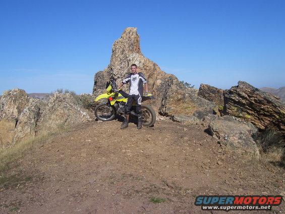 Man in riding gear stands with yellow dirt bike on a rocky hill under a clear blue sky. Supermotors.net logo in corner