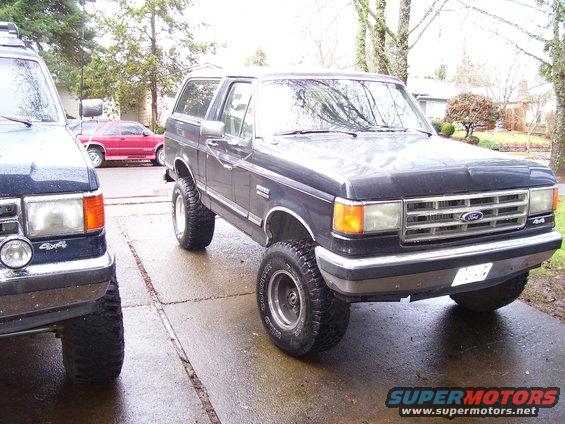 Dark blue Ford Bronco with lifted suspension and off-road tires, parked on a wet driveway next to another SUV
