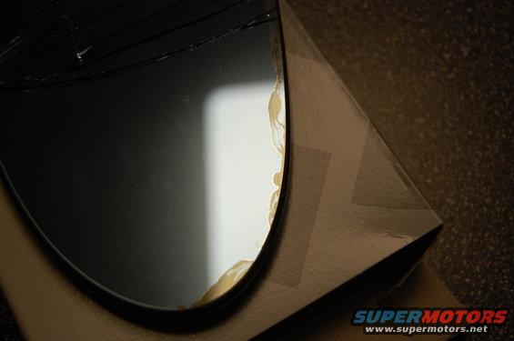 Broken mirror with yellowed adhesive on edge, resting on cardboard packaging