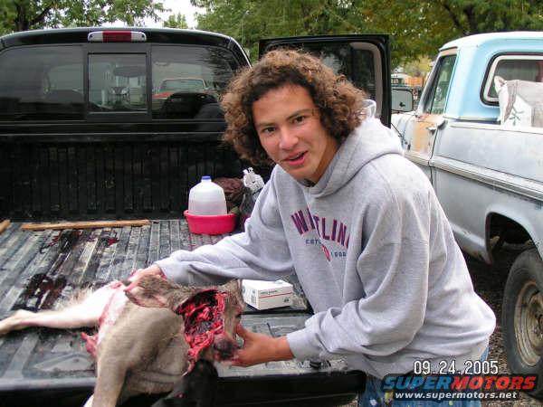Man in gray hoodie with deer carcass in black truck bed next to a light blue pickup truck