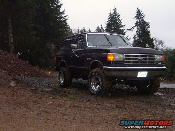 1988 Ford Bronco parked on a muddy, wooded hill with trees in the background