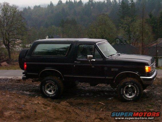 Black Ford Bronco parked on muddy ground with trees and houses in background