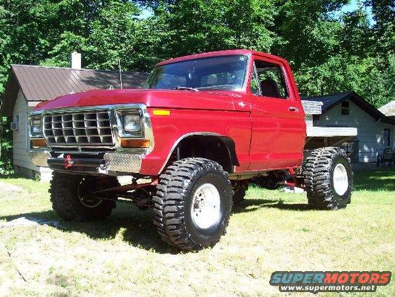 Red pickup truck with large tires and a flatbed, parked in a grassy yard with houses in the background