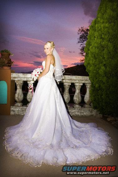 Bride in a white wedding dress with a veil and bouquet stands on a balcony at sunset